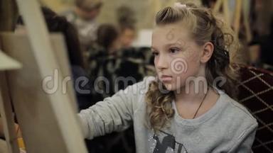 坐在画架上的漂亮少女在一所艺术学校画一幅画。 儿童专业艺术学校